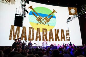 A band stands in front of a large electronic sign with a logo for the Madaraka Festival. The venue is full of people.