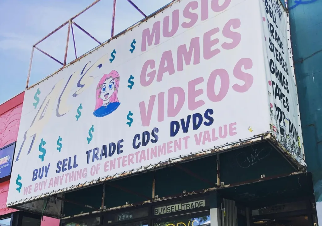 Exterior shot of Al's Music, Games, & Videos with its bright colored sign. A shopkeeper stands in front of the store.
