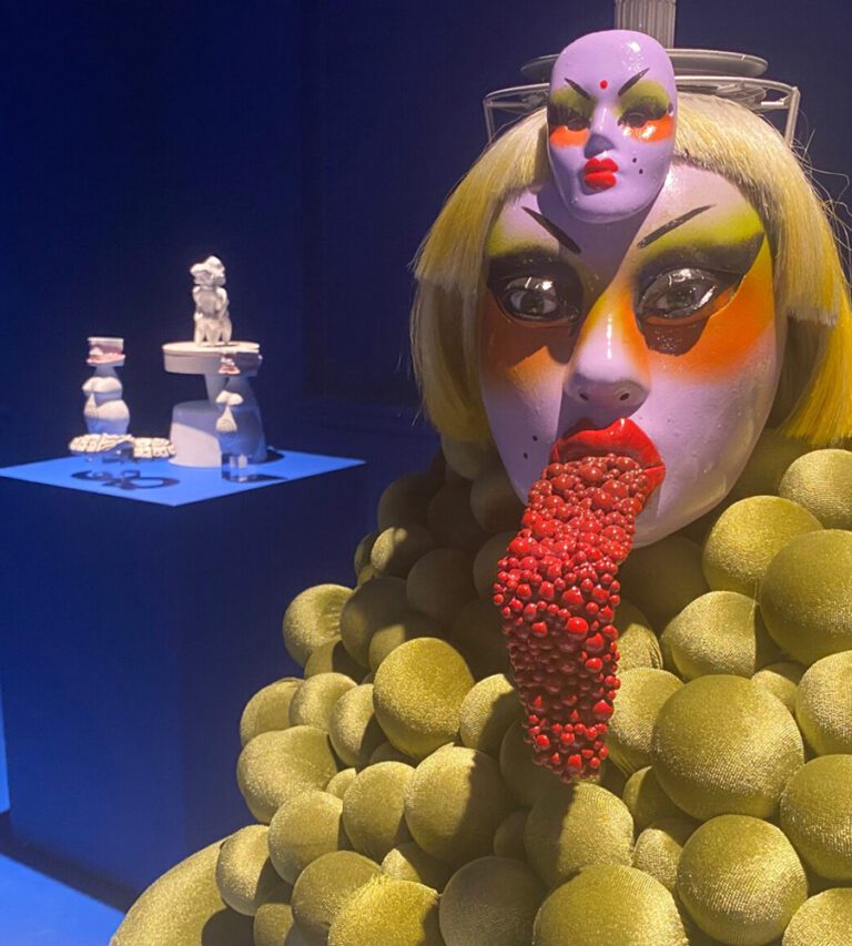 A colorful mannequin with a giant bumpy red tongue in front of smaller sculptures