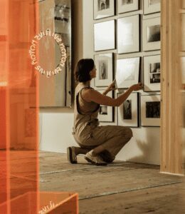 A person kneels down to hang a framed photo inside a wooden, airy, well-lit space.