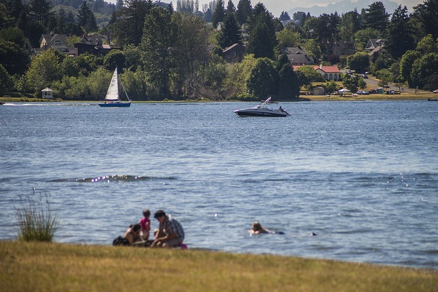A family lays on the grass in Seward Park in front of the water as a sailboat and motor boat drift across the water