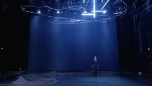 A shot from a trailer for Where We Belong, featuring a solo performer on a bare stage with lights floating above her