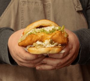 A sandwich with Beer-Battered Alaskan Pollock, pickle-ly tartar sauce, shredded lettuce, American cheese on a Lil Woody’s bun