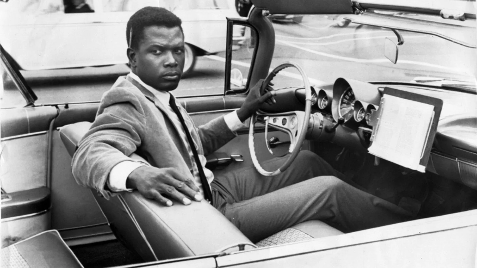A screencap from The Slender Thread. Sidney Poitier sits in a classic convertible looking over his shoulder