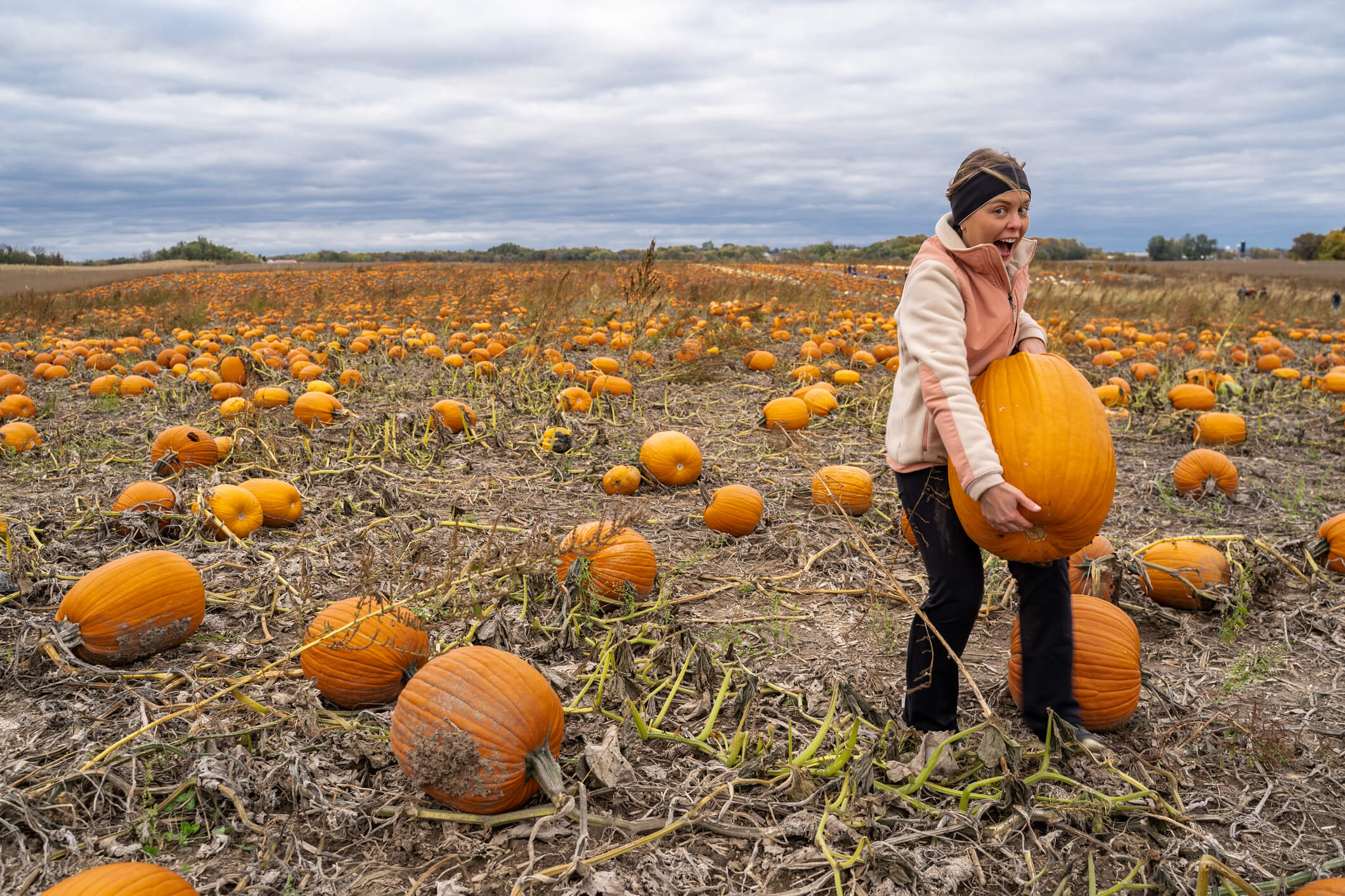 Adult woman (30s) attempts and struggles to lift and to pick up a giant pumpkin from a pumpkin patch. Smiling and laughing and having fun
