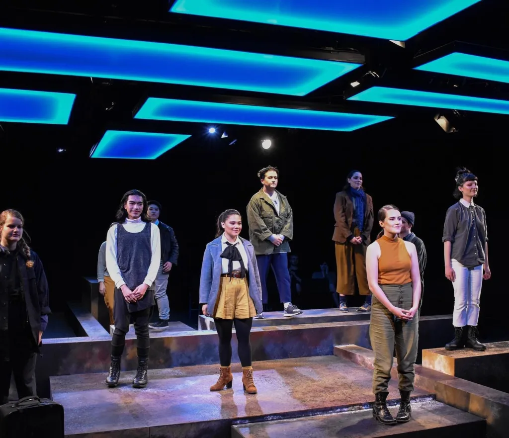 A photo of the cast from a Seattle University theater production posing on stage underneath square, blue lights