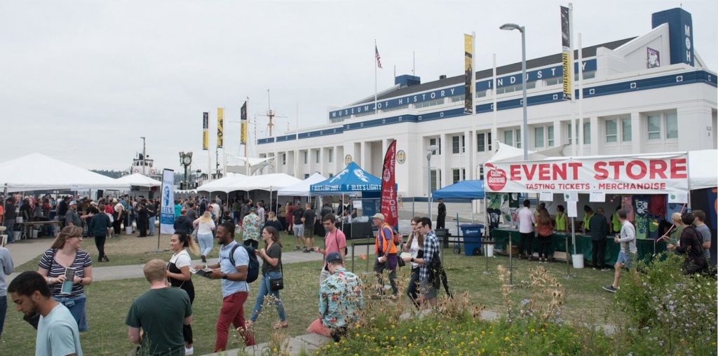 A crowd of people at a Seattle Cider Summit at MOHAI with a large building and lots of flags in the background