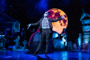 A photo of Jack Skellington with arms outstretched, centerstage. Sally leans against a prop in the background.