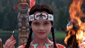 Christina Ricci as Wednesday Adams lights a match, a giant fire burns behind her. She wears an Indigenous outfit.