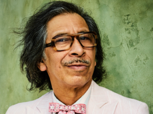 An image of Kid Congo Powers looking directly into the camera, wearing brown-rimmed glasses and a pink bowtie