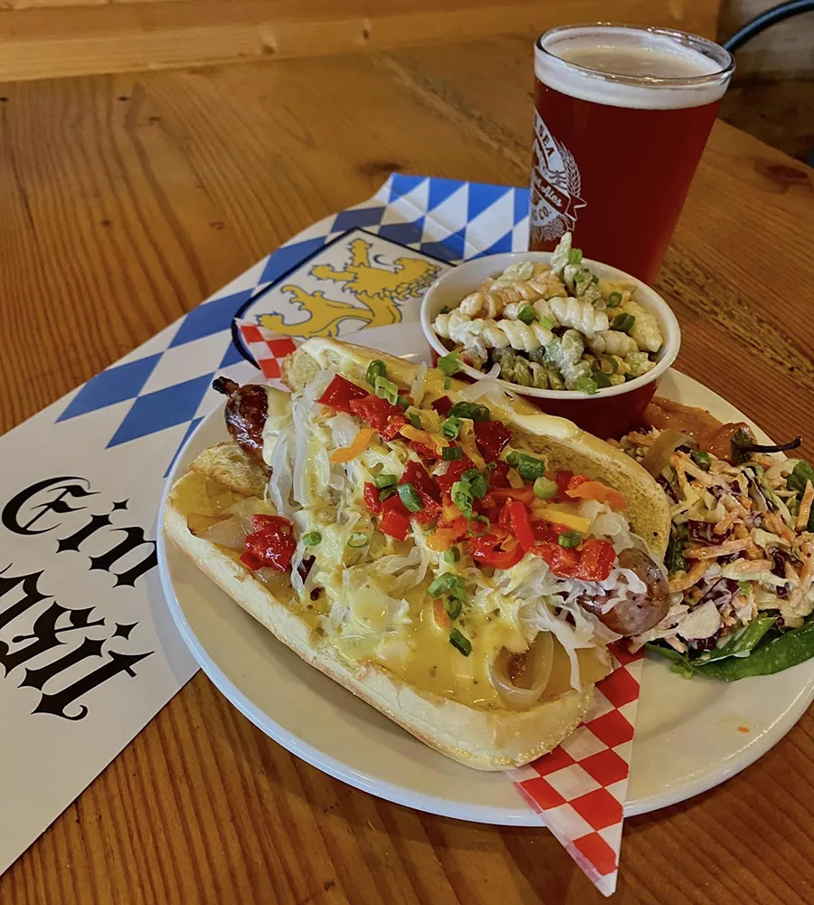 A bratz dog with a mound of sauerkraut, bell peppers, and green onions. Served with cabbage slaw, pasta salad and a glass of beer on a wooden table