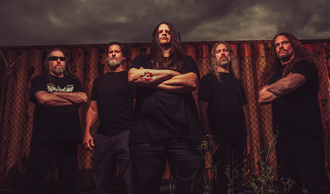The band Cannibal Corpse posing in front of a shipping container underneath a stormy, gray sky.