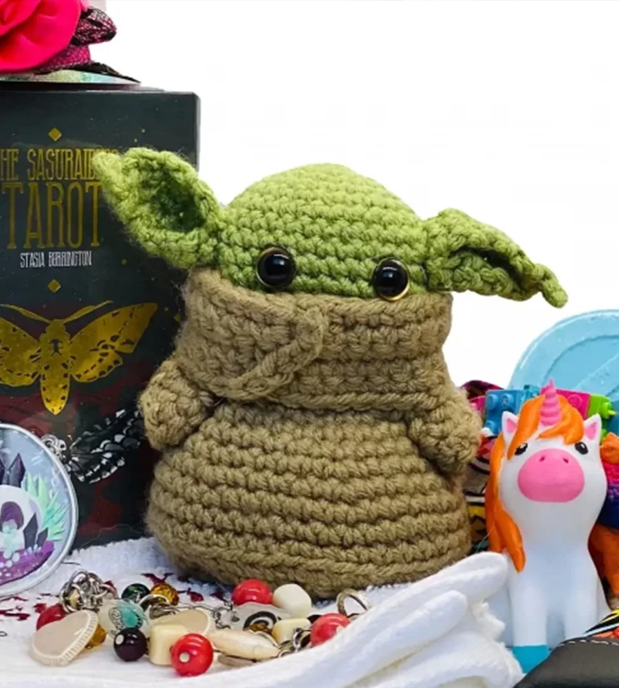 A little knitted baby yoda sits at the center of an array of geeky gifts, like a unicorn and tarot deck.