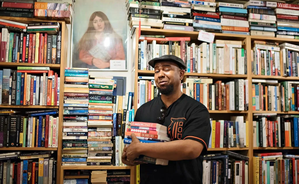 Owner of BLMF Literary Saloon stands in front of a stack of books and holds one in the crook of his arm
