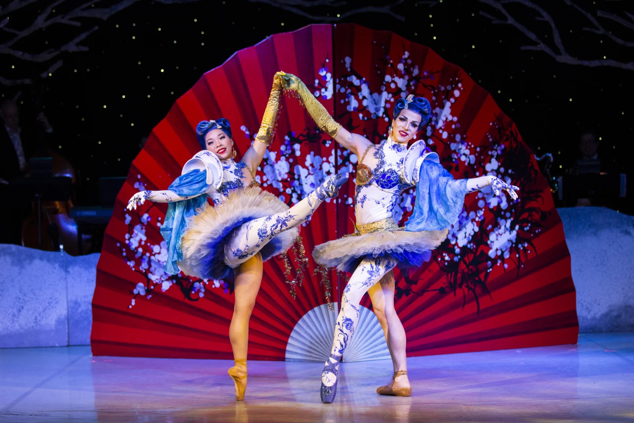 Two dancers in tutus hold hands while a giant fan is displayed behind them