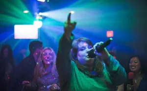 A woman singing through a microphone points up towards the ceiling in a crowded blue-green lit bar