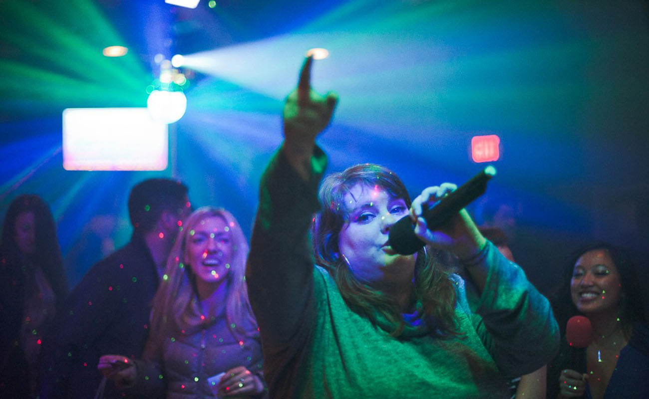 A woman singing through a microphone points up towards the ceiling in a crowded blue-green lit bar