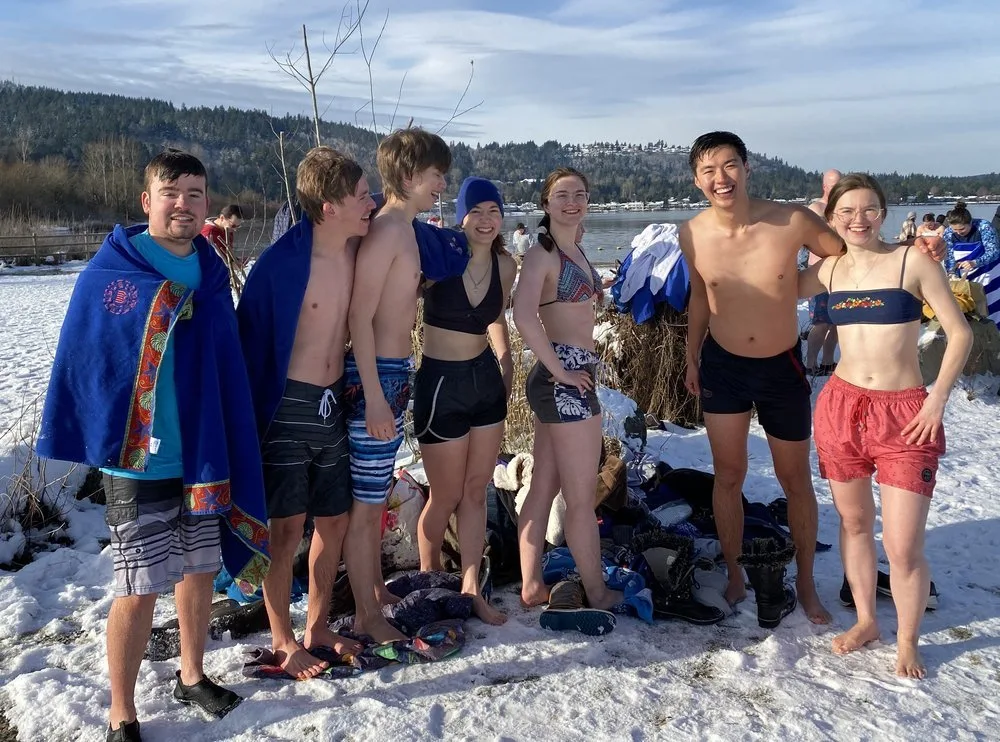 A group of smiling people in bathing suits pose for a photo on a snow-covered beach after a polar bear plunge.
