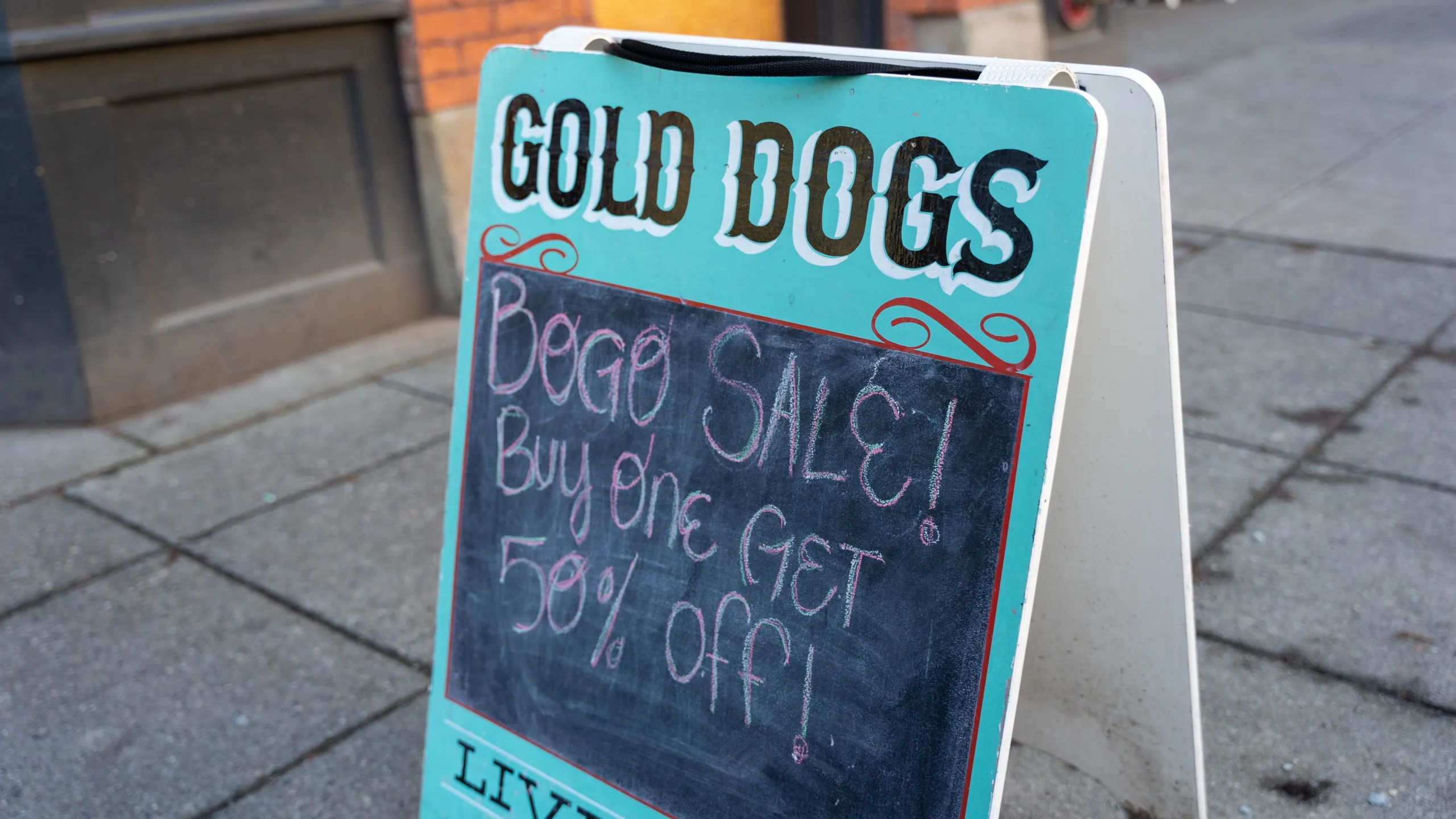 A turquoise sign outside of Gold Dogs on Ballard Ave reads "GOLD DOGS" and lists a "BOGO SALE" in chalk