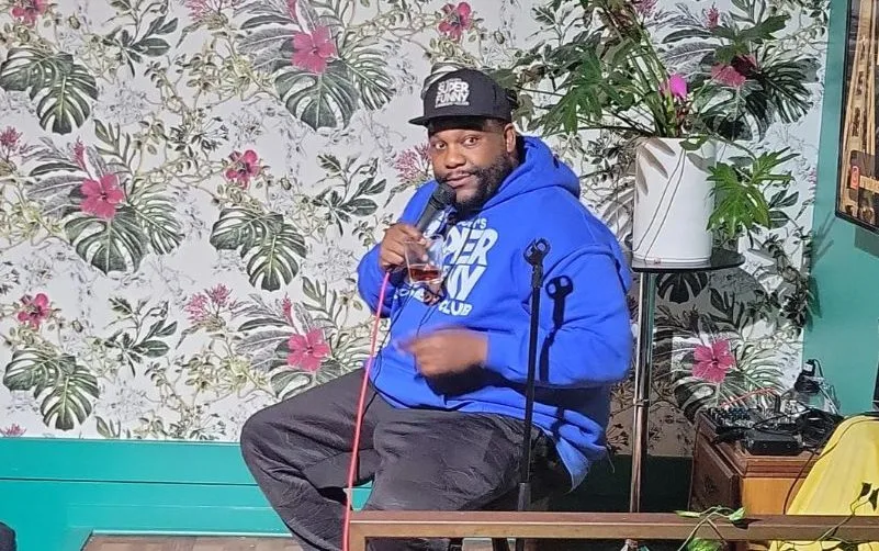 A comedian sits inside a colorful, tropical room with a drink in hand