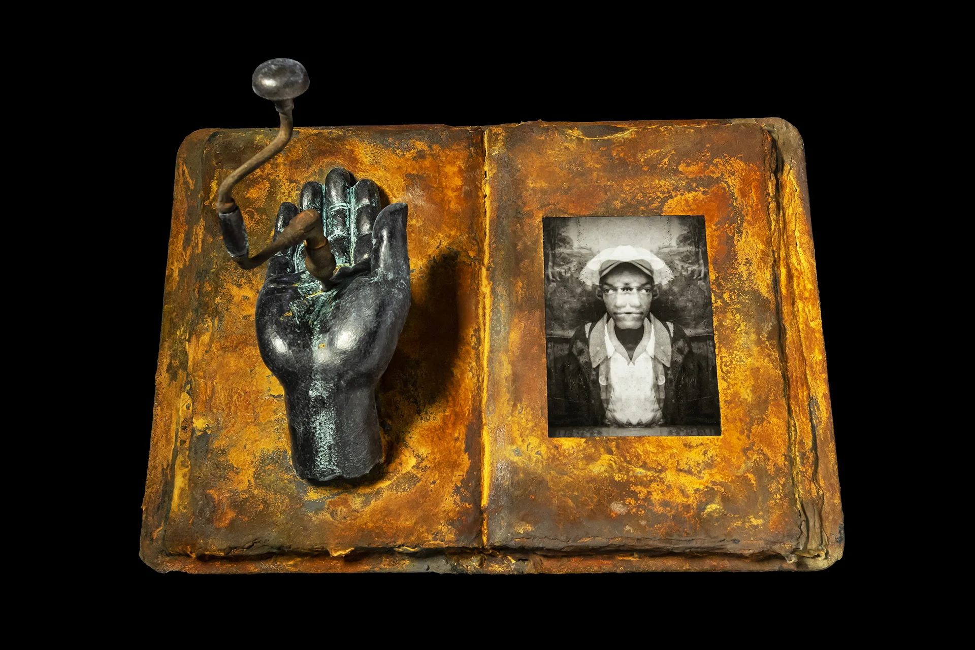 A metal, bronze book has a sculpture with a hand rising out its leftside and an antique, blurred photo of a man on the right side