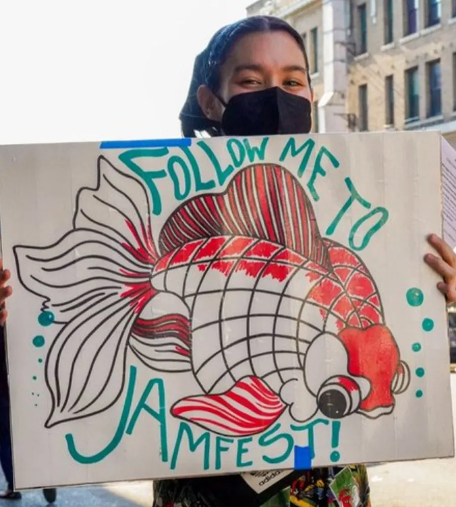 A volunteer holds up a sign that reads "FOLLOW ME TO JAMES ST!" with a red koi fish drawn in the center