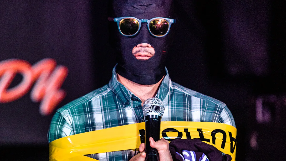A performer performs at Safeword with a mask over their face, glasses, and their hands tied up by caution tape. A microphone is held up by their mouth.