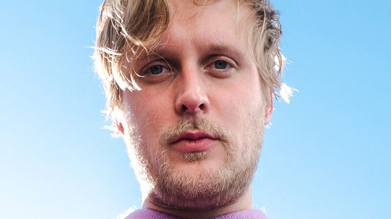 John Early in a lilac shirt against a pale blue sky