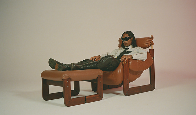 Steve Lacey reclines on a brown leather chair and ottoman against a hazy, taupe background.