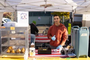 A person stands behind a Seattle Samosa stand at a Farmers Market in Seattle flashing a thumbs up.