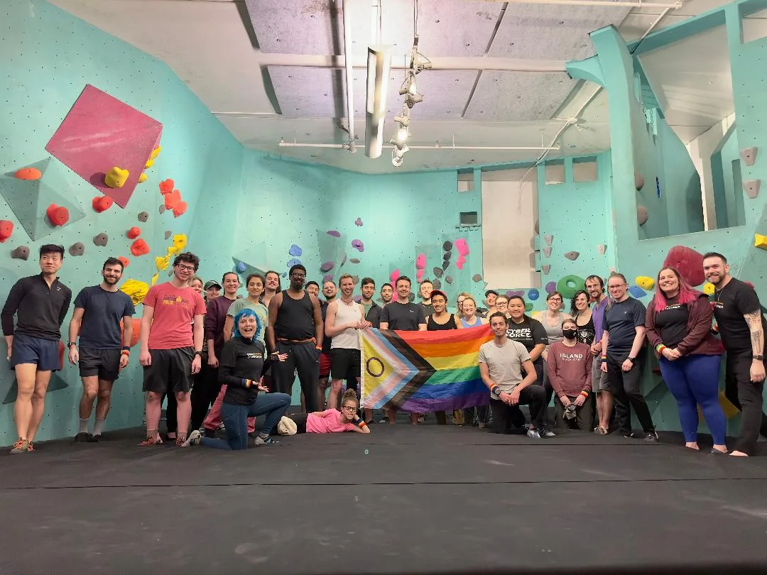 A large group of climbers stand in front of a baby blue climbing wall while holding a queer flag in the center.