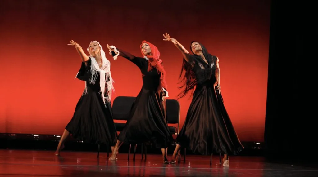 Three women in a line in long, black robes reach an arm towards something offstage. 