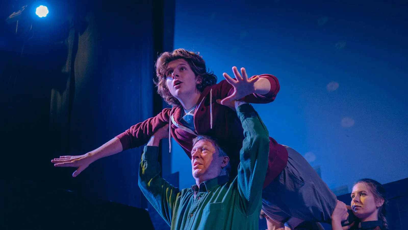 A production photo from the play The Curious Incident of the Dog in the Night-Time. The main character, Christopher is lifted into the air, Superman-style by his dad and a woman.
