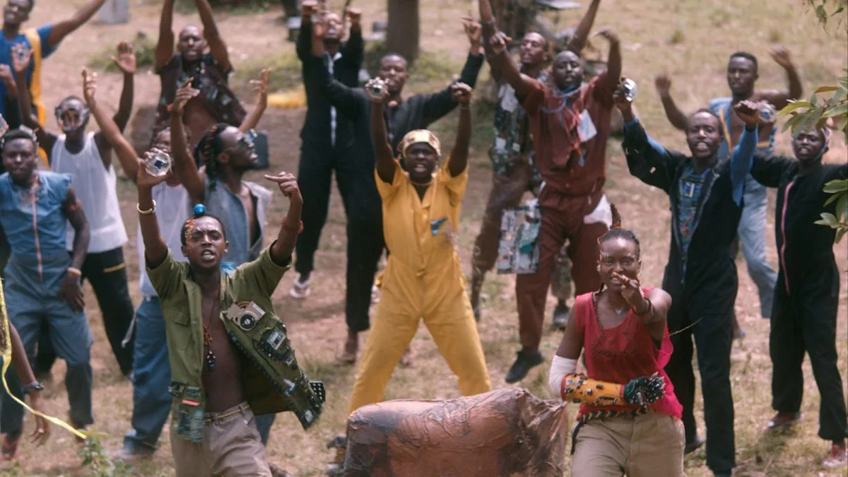 A screencap from the movie Neptune Frost. A group of people stand in a field, facing the viewer mid song