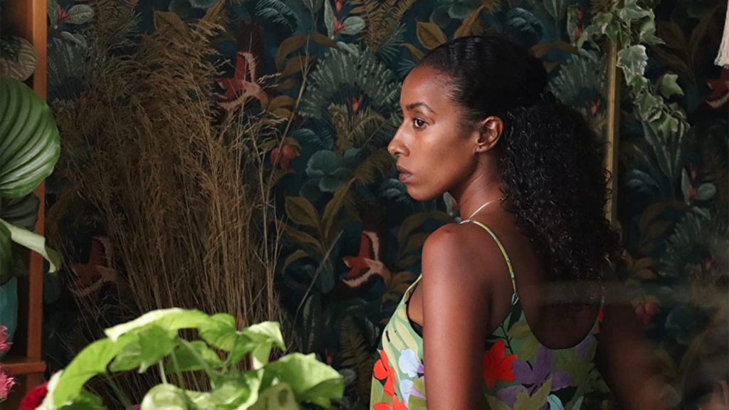 A screencap from the film "America." Actress Oshrat Ingadashet who plays Iris in the film stands against a dark green botanical wallpaper in a plant-filled room and looks off to the side.