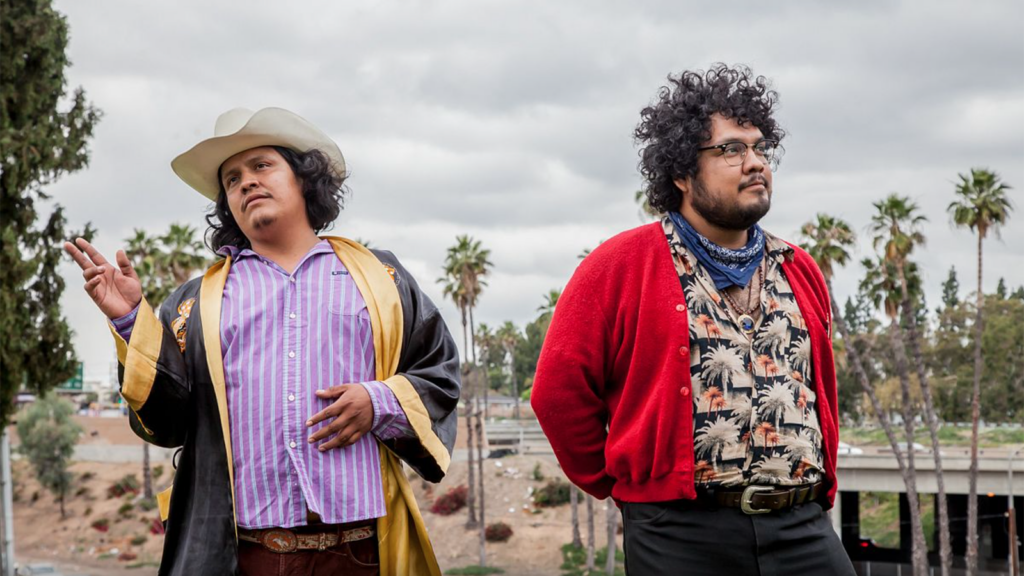 Brothers David and Rene Pacheco of Tropa Magica stand off the side of a road under a cloudy sky in colorful outfits.