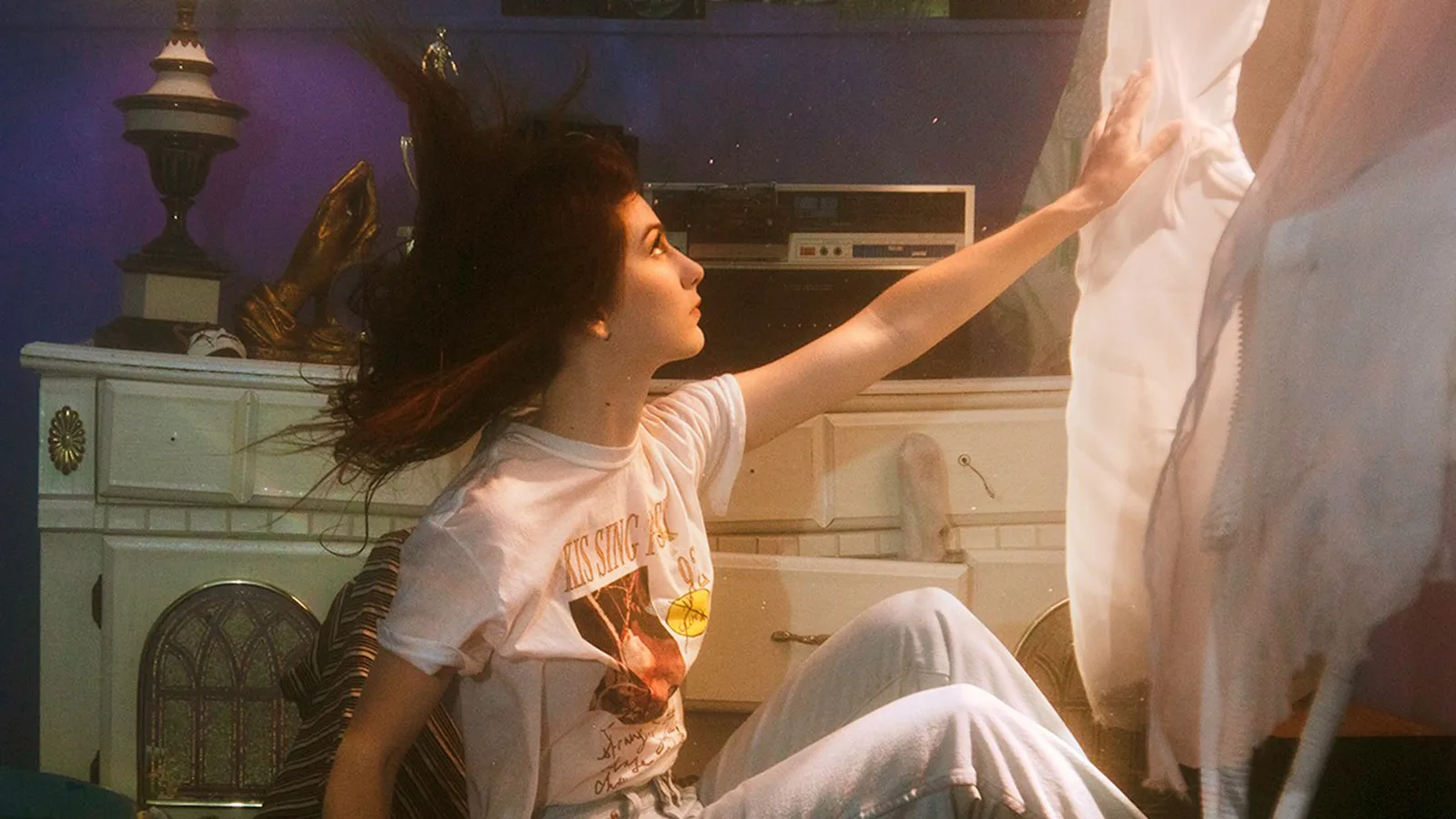 The artist Weyes Blood in an underwater photoshoot reaching towards a bright light off to the right.