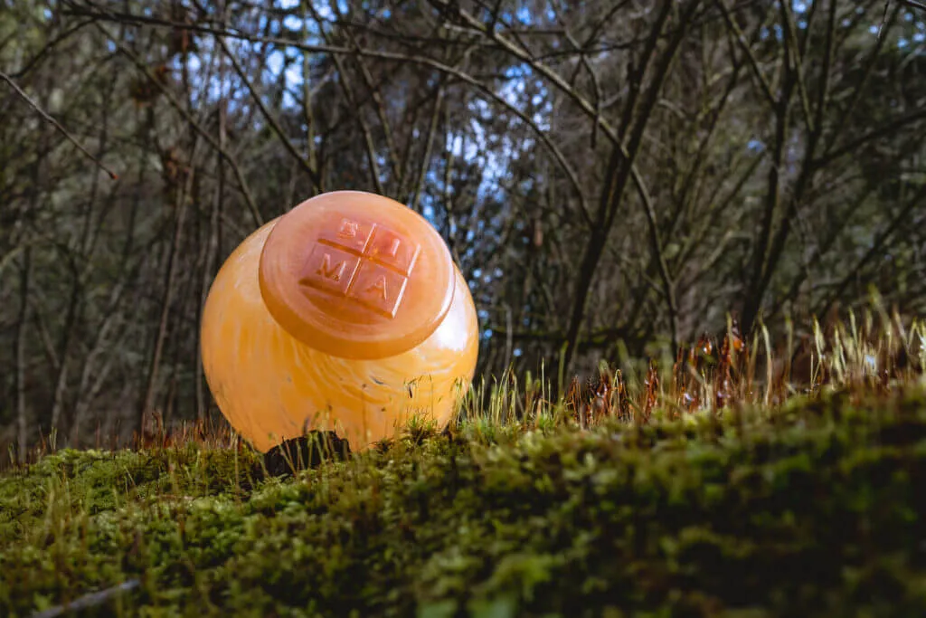 A shining orange glass orb with the Bainbridge Island Museum of Art's logo stamped onto it rests on a grassy hill with sunlight against it.
