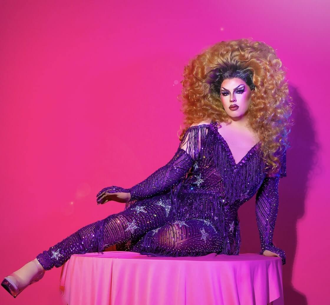 A drag queen with big hair and big beaded fringe against a pink background