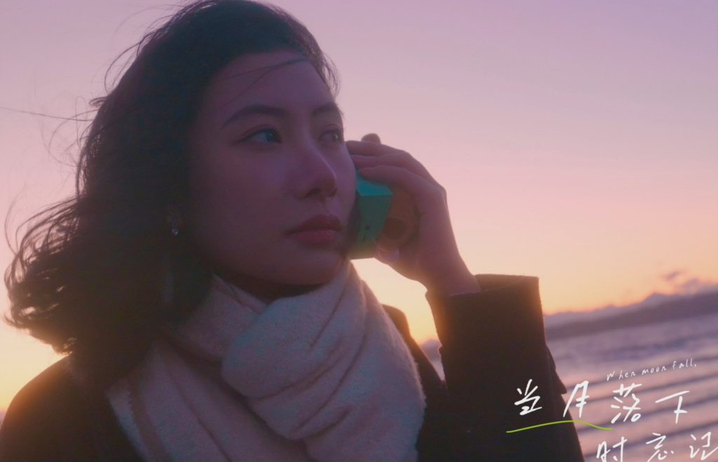 A woman looks to the left, holding a phone to her ear as the wind blows her hair. She's by the water at sunset.