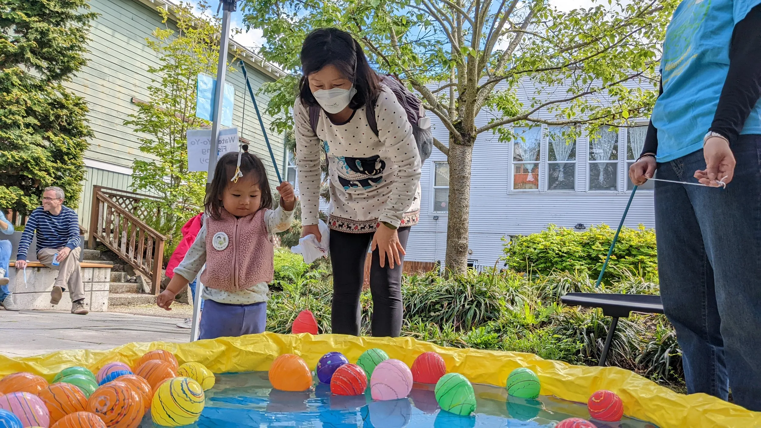 A mother and child stand in front of a kiddie pool filled with water and floating mini balloons.