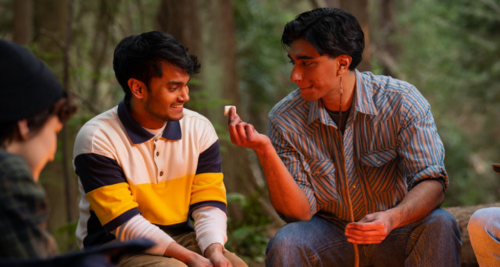 Two young men sit together by a campfire. They smile at each other while one offers the other a marshmallow