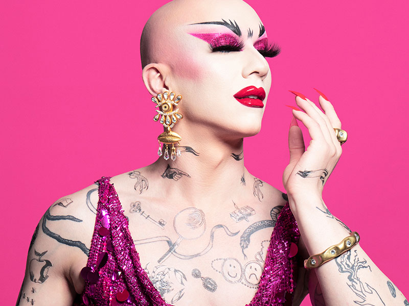 Drag queen Sasha Velour looks to the right with her eyes closed and hand raised. She stands in front of a pink background and wears a pink dress and pink lipstick and eye shadow.