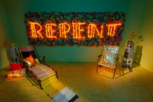 A colorful room with the words REPENT spelled in neon in the background. In the foreground, two chairs invite people to sit on them. Meryl Pataky (American, born 1983) with textile collaboration by Allie Felton. A Modern Guilt (installation view), 2020. Neon and mixed media; dimensions variable. Courtesy of artist. Photo by Deb Leal.