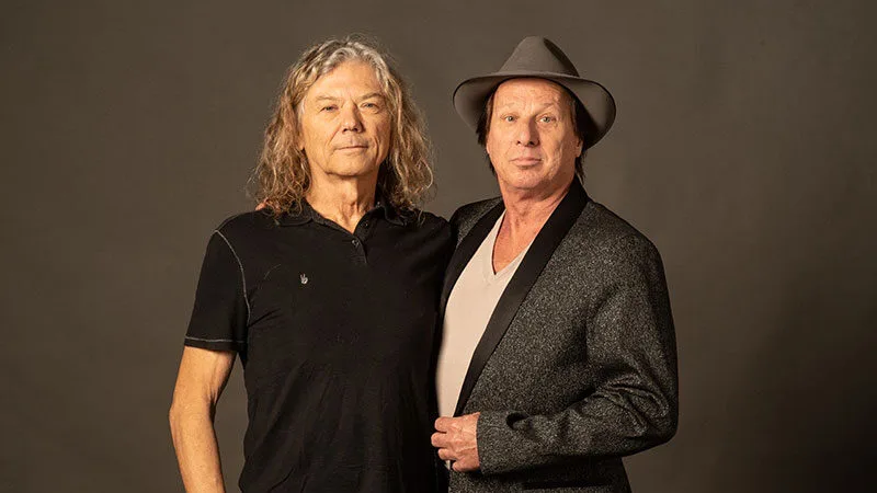 Musicians Jerry Harrison and Adrian Belew pose together against a brown background.