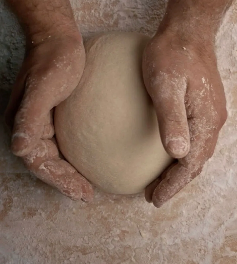 A screencap from the movie Stella. Two hands are cupped around a ball of pizza dough on a floured counter as they knead.
