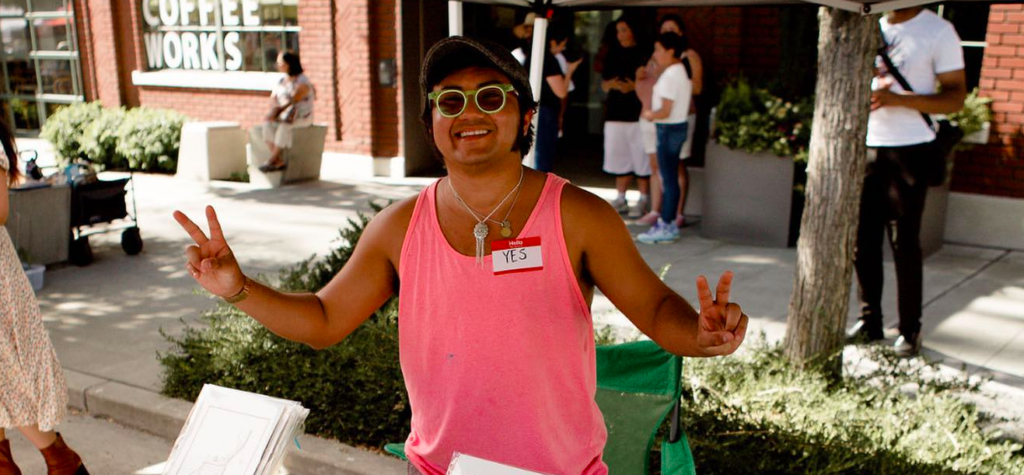 A person in a salmon tank top smiles at a camera while flashing peace signs and standing above a table at a market.