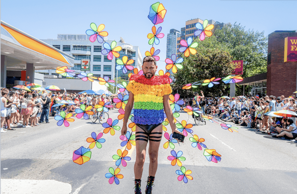 A person in a bright rainbow outfit walks down the middle of the street in a Pride parade