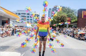 A person in a bright rainbow outfit walks down the middle of the street in a Pride parade