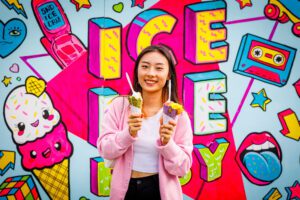 A woman stands in front of a colorful ice cream mural holding two ice cream cones.